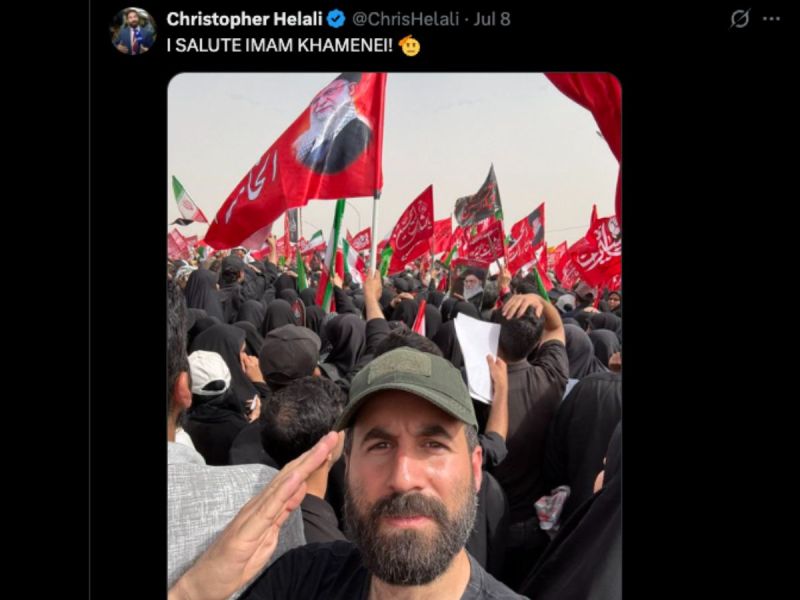 A man salutes while taking a selfie in a crowd holding red and black flags with Arabic writing at an outdoor event.