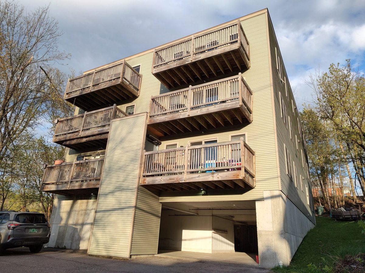 A three-story apartment building with multiple wooden balconies and a partially open ground-level parking area, surrounded by trees.