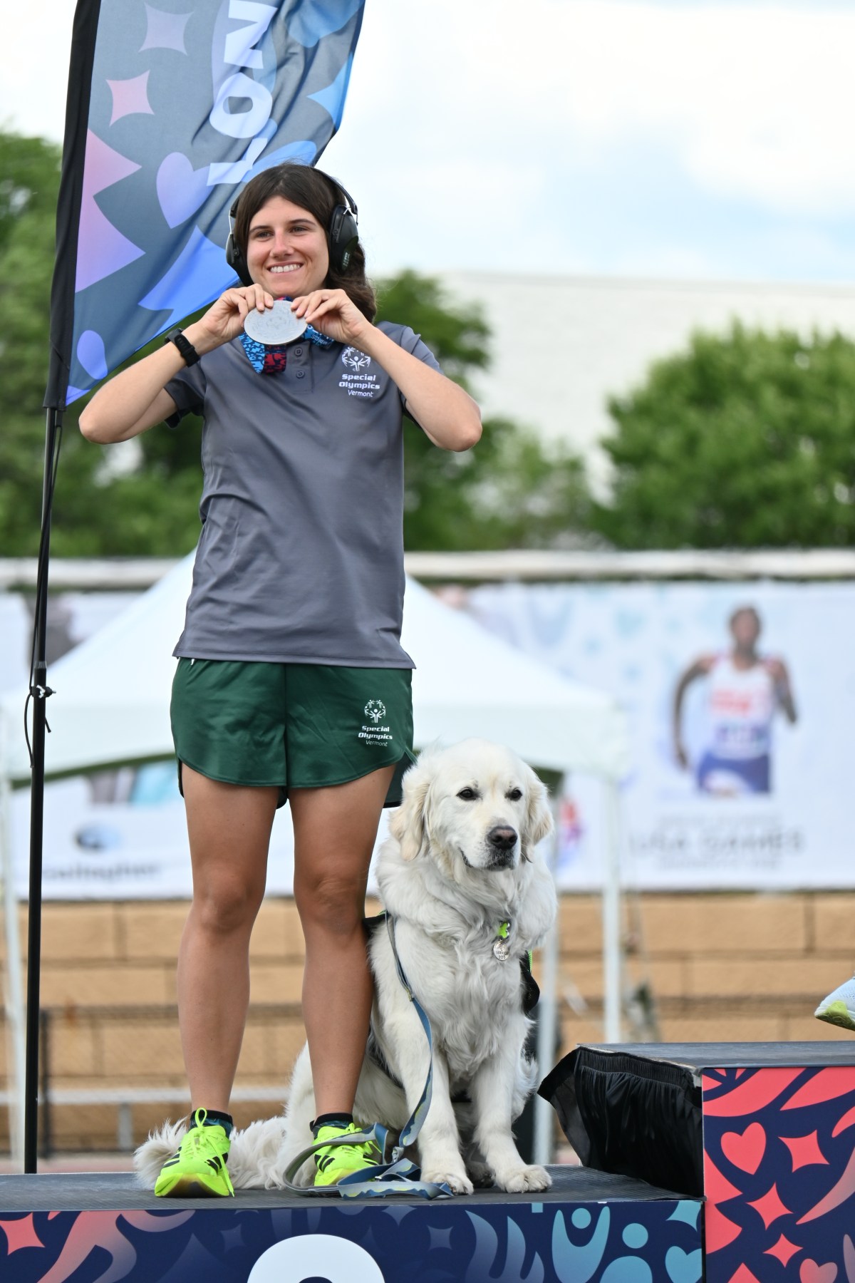 A woman in sportswear stands on a podium beside a golden retriever, signing with her hands. Event banners and a blurred runner's poster are visible in the background.