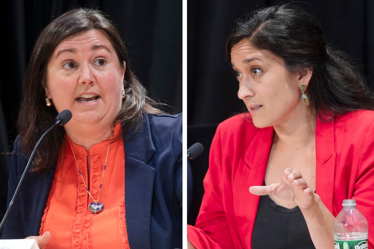 Two women sit at microphones, speaking and gesturing during a discussion. Both wear blazers; one is in navy and orange, the other in red, with a water bottle visible in front of the second woman.