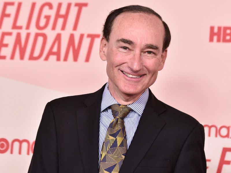 A man in a suit and patterned tie smiles at a red carpet event with “Flight Attendant” and “HBO” visible on the backdrop behind him.