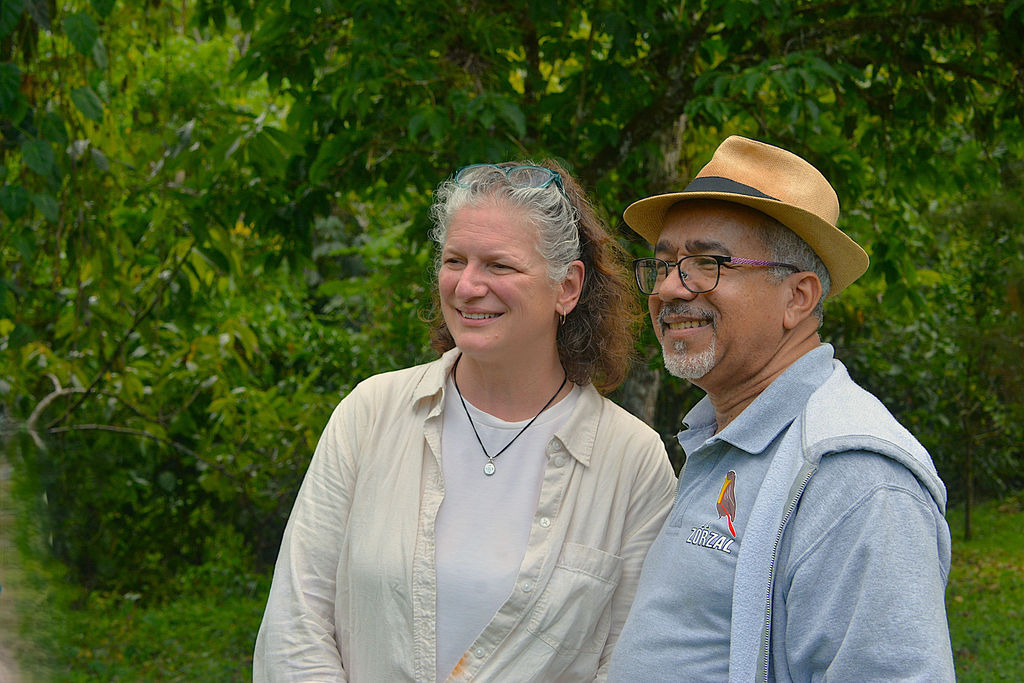 Two adults stand outside in front of greenery, smiling and posing for a photo. One wears a hat and glasses; the other has curly hair and a light shirt.