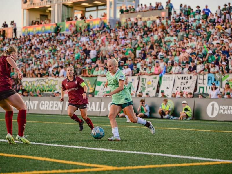 A soccer player in a green uniform dribbles the ball while two players in maroon chase, with a large crowd watching from the stadium in the background.