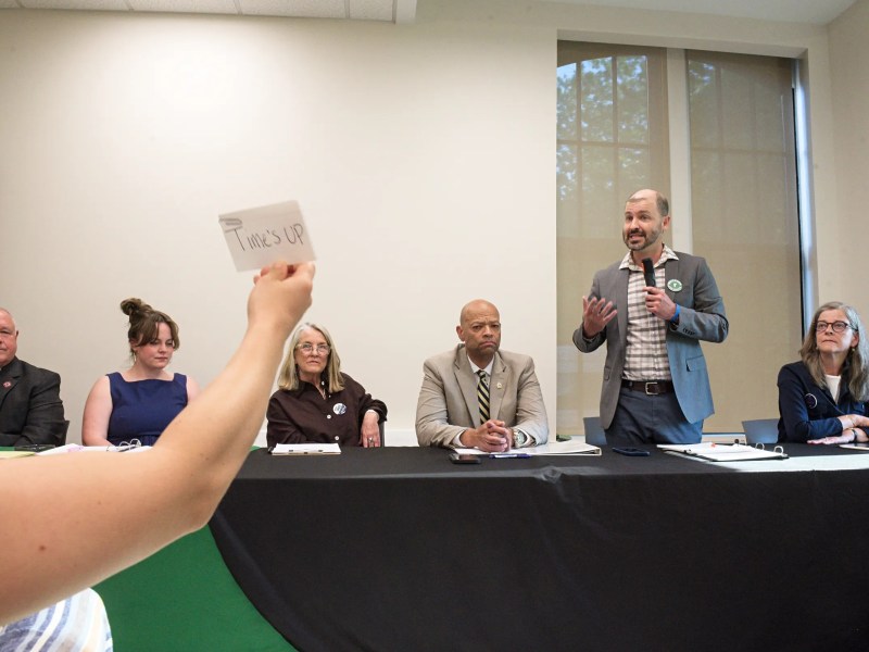A person holds up a "TIME'S UP" card while a man speaks at a panel discussion with five seated panelists at a table.