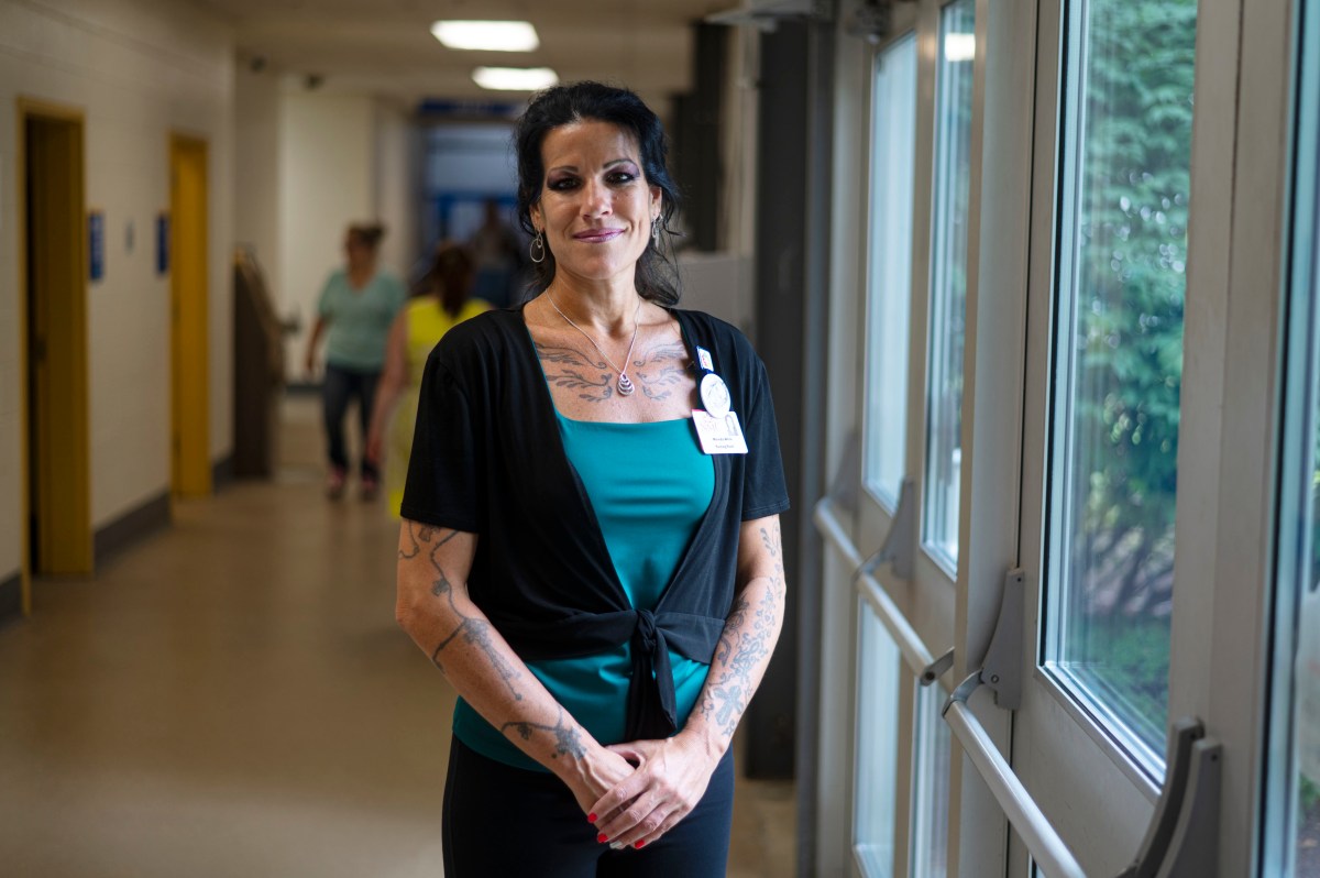 A woman with tattoos and a staff badge stands by a window in a hallway, facing the camera. People are visible in the background.