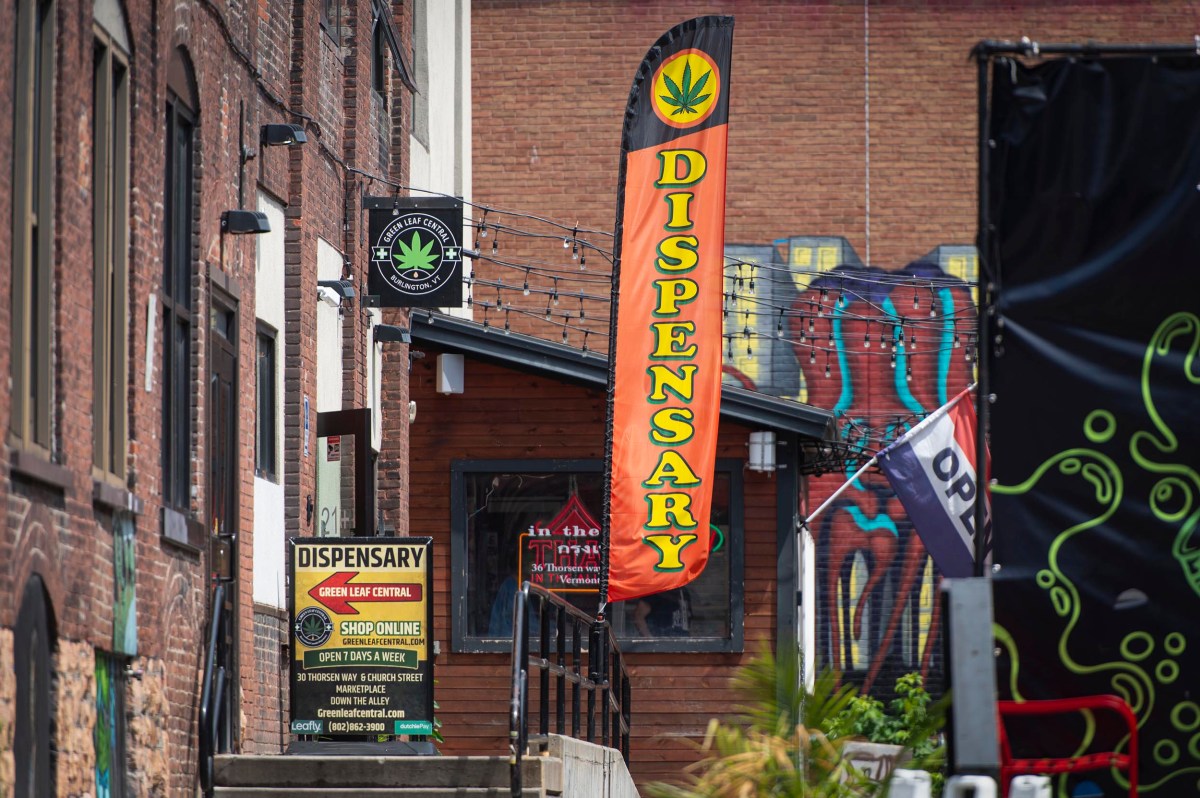 A marijuana dispensary with a large "Dispensary" flag, exterior signage, and an "Open" flag is shown in an urban area with brick buildings and graffiti.