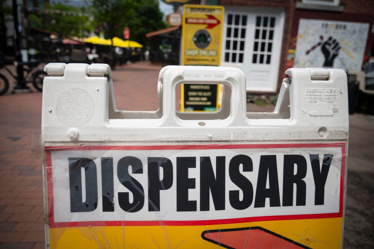 A sign with large black letters reading "Dispensary" stands on a brick sidewalk in front of storefronts and outdoor seating.