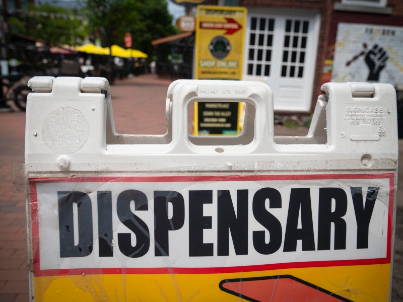 A sign with large black letters reading "Dispensary" stands on a brick sidewalk in front of storefronts and outdoor seating.
