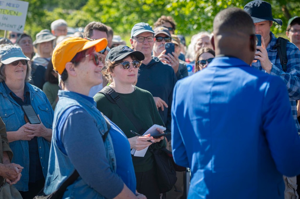 A group of people gathers outdoors, listening to a person speaking while some attendees take notes or record with their phones.