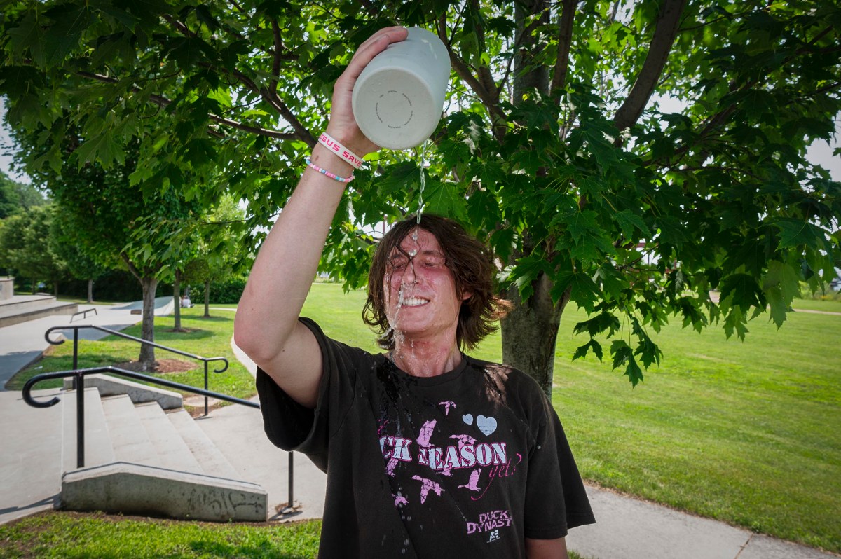 A person standing outdoors pours a white liquid over their head, with liquid splashing onto their face and shirt. A tree and grassy area are visible in the background.