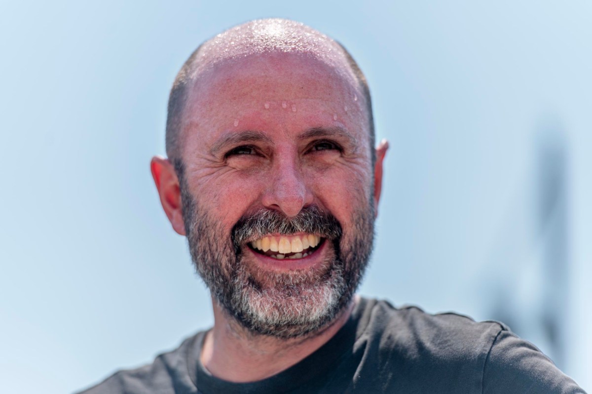 A smiling man with a beard and short hair, wearing a black t-shirt, is pictured outdoors on a sunny day with visible sweat on his forehead.