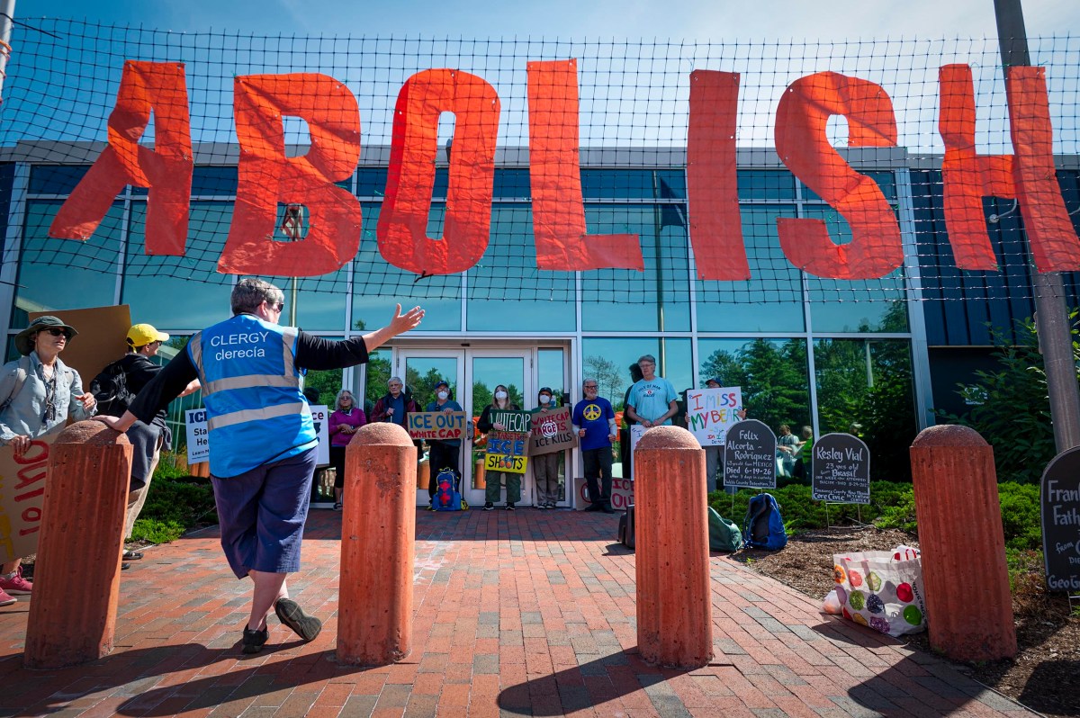 Protesters stand in front of a building with a large "ABOLISH" sign overhead, holding various signs. One person in a blue "CLERGY" vest gestures toward the group.