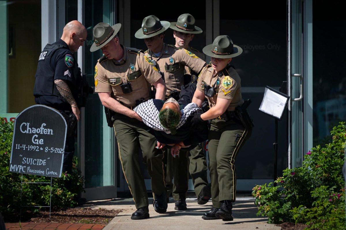 Four uniformed officers carry a person out of a building entrance while another officer stands nearby. A sign with writing is visible on the left side of the image.