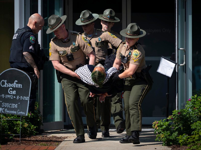 Four uniformed officers carry a person out of a building entrance while another officer stands nearby. A sign with writing is visible on the left side of the image.