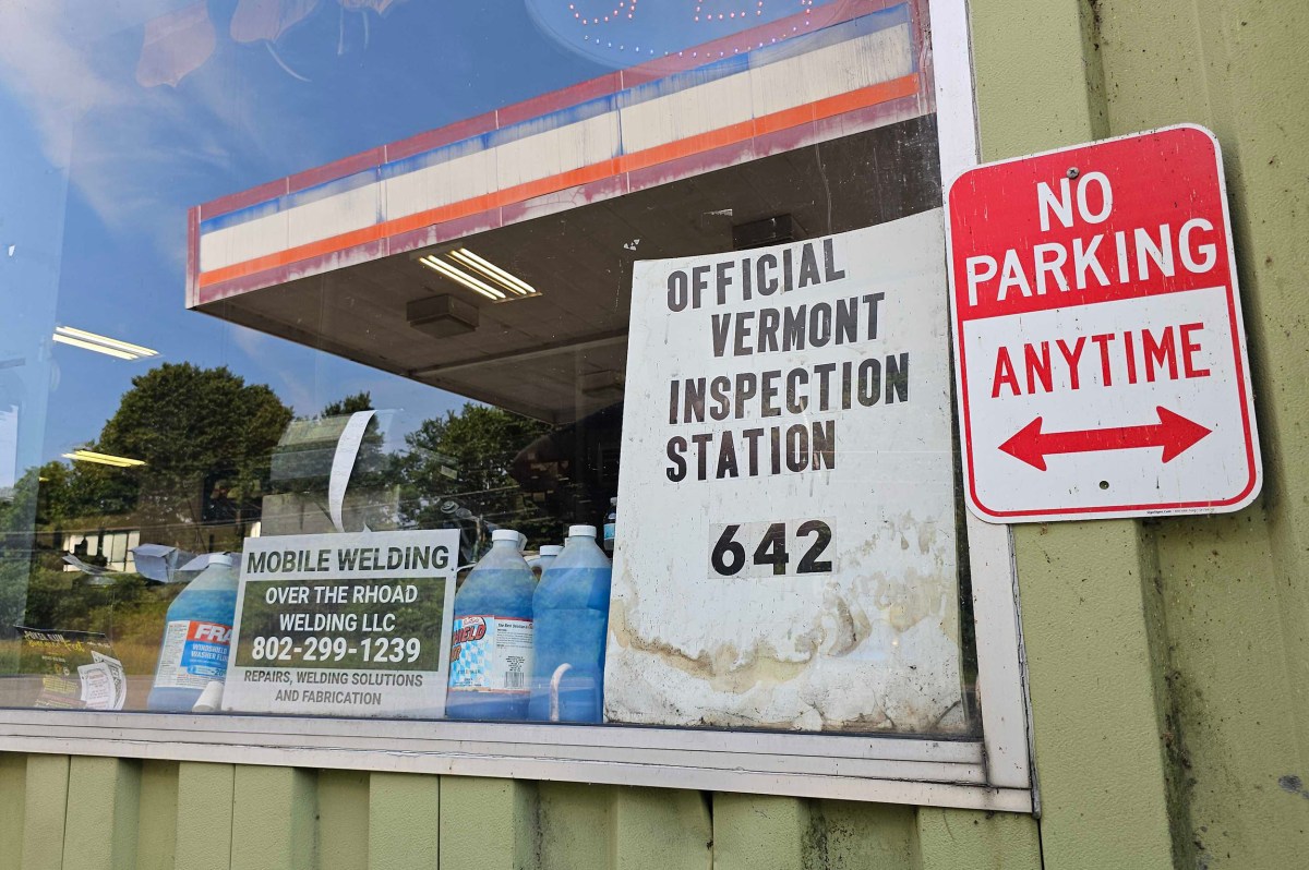 A window displays signs for an official Vermont inspection station, a mobile welding service, and a red "No Parking Anytime" sign. Blue jugs and trees are visible through the glass.