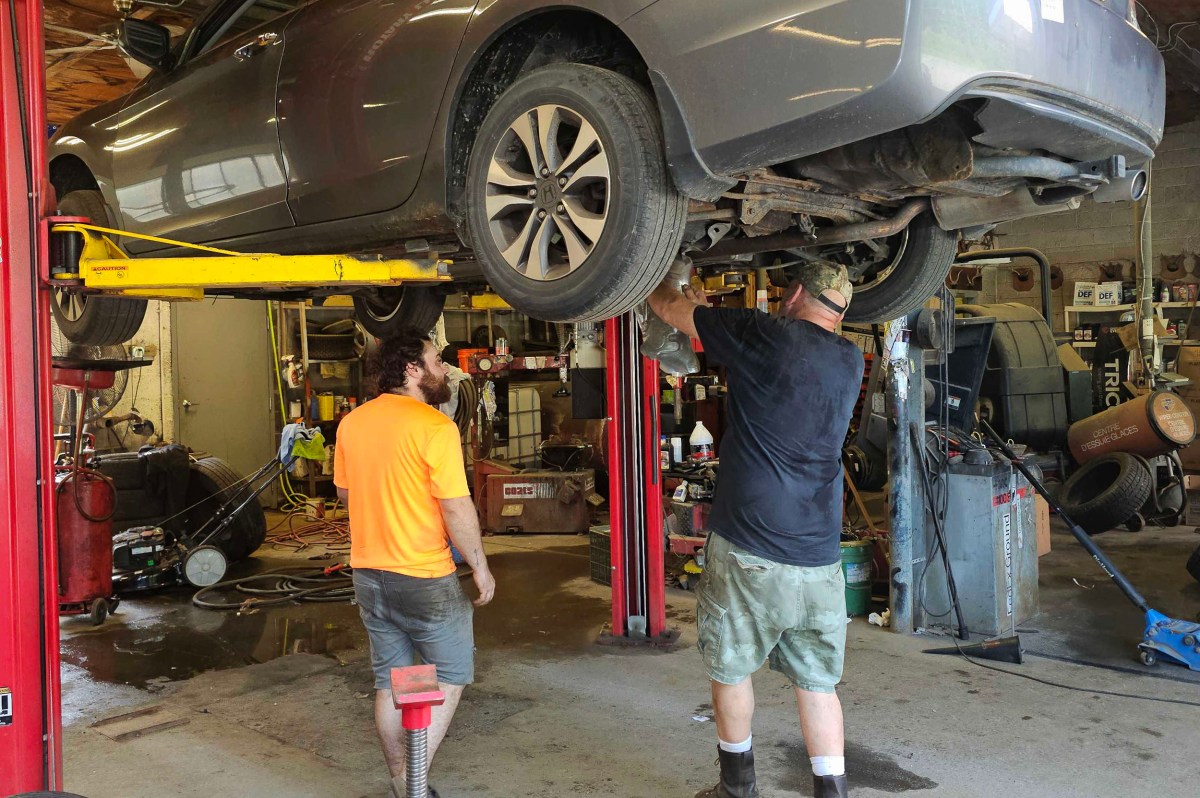 Two mechanics work on a car lifted on a hydraulic lift inside an auto repair garage, with tools and equipment visible in the background.