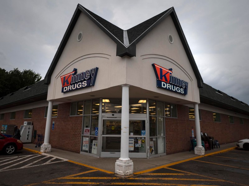 Exterior view of a Kinney Drugs store on a cloudy day, with glass entrance doors, parking spaces, and visible signs above the doorway.