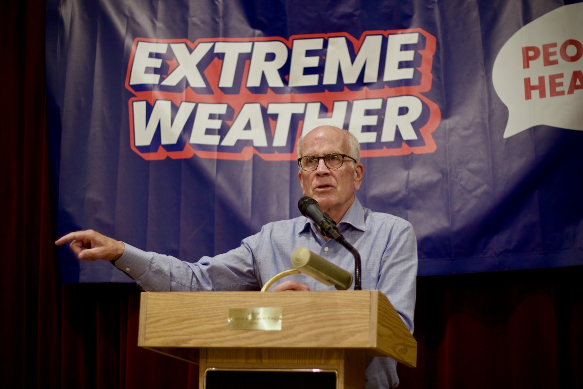 An older man speaks at a podium, gesturing with his left hand. Behind him is a banner that reads "EXTREME WEATHER" in bold letters.