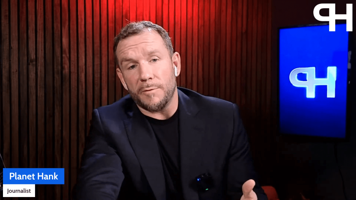 A man in a dark blazer speaks on camera in a studio with wood paneling, a blue screen, and "Planet Hank Journalist" text label in the corner.