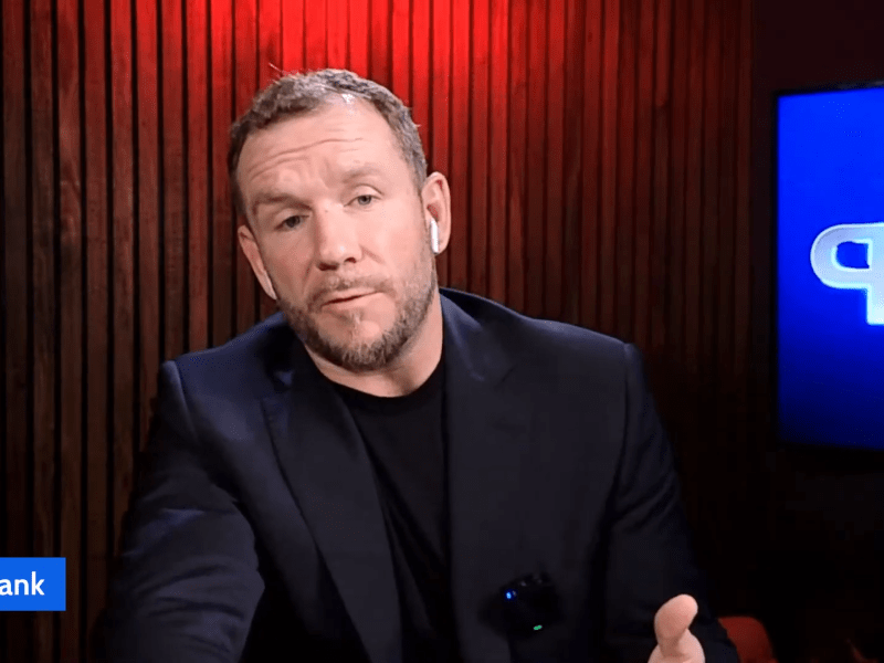 A man in a dark blazer speaks on camera in a studio with wood paneling, a blue screen, and "Planet Hank Journalist" text label in the corner.