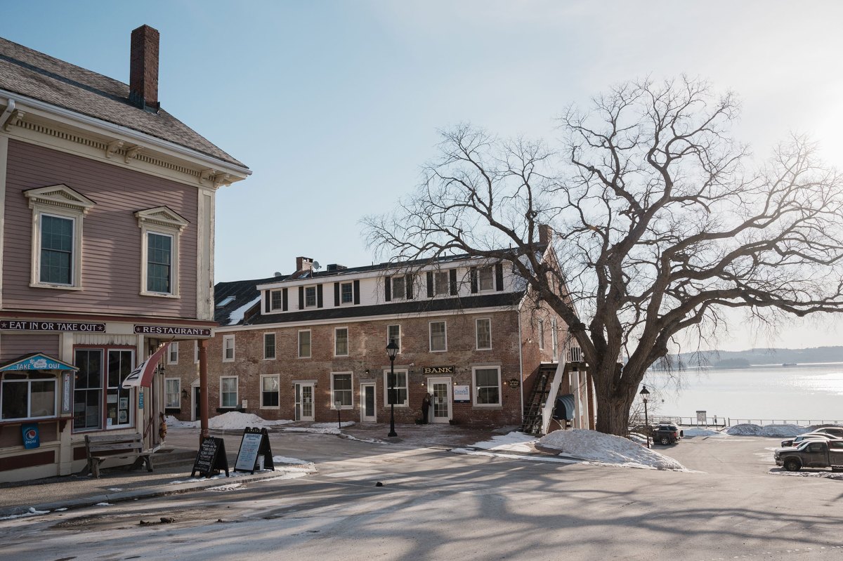 Small historic town square with brick buildings, a bank, a restaurant, bare tree, patches of snow, and a river in the background under clear sky.