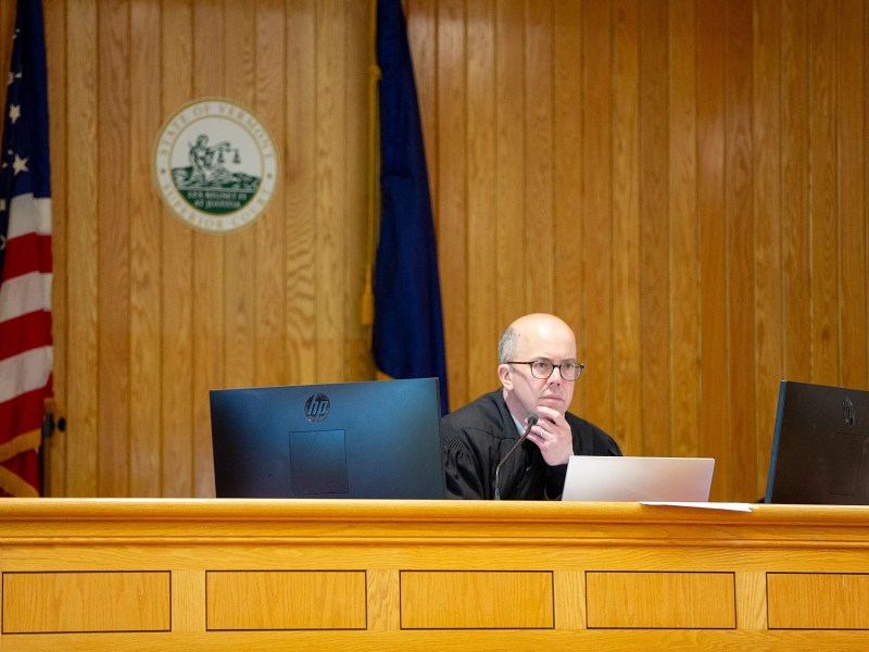 A judge sits at a wooden bench between two monitors in a courtroom, with an American flag and a seal on the wood-paneled wall behind.