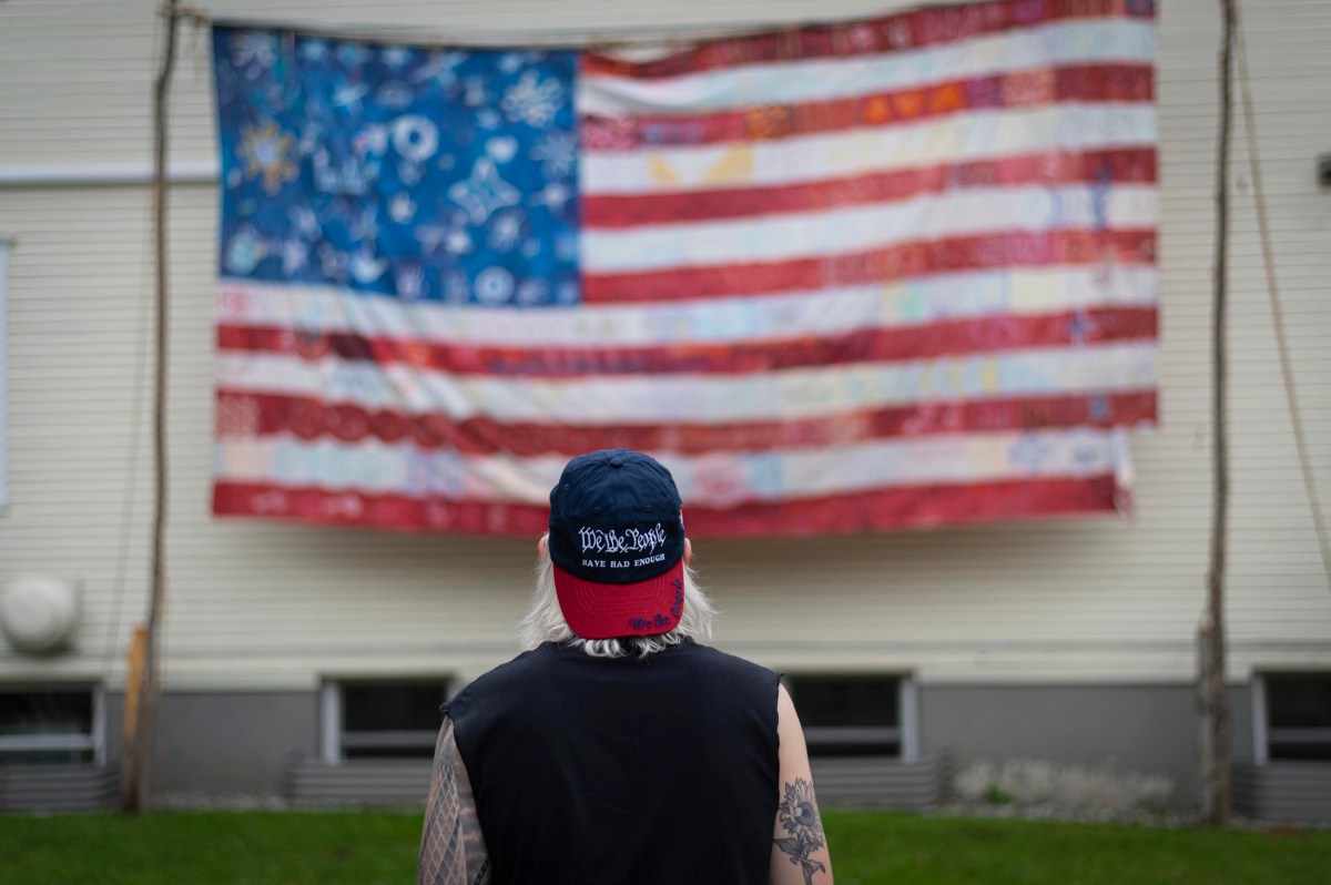 A person with long hair and tattoos, wearing a blue and red cap, stands facing a large hand-made American flag hanging on a building.