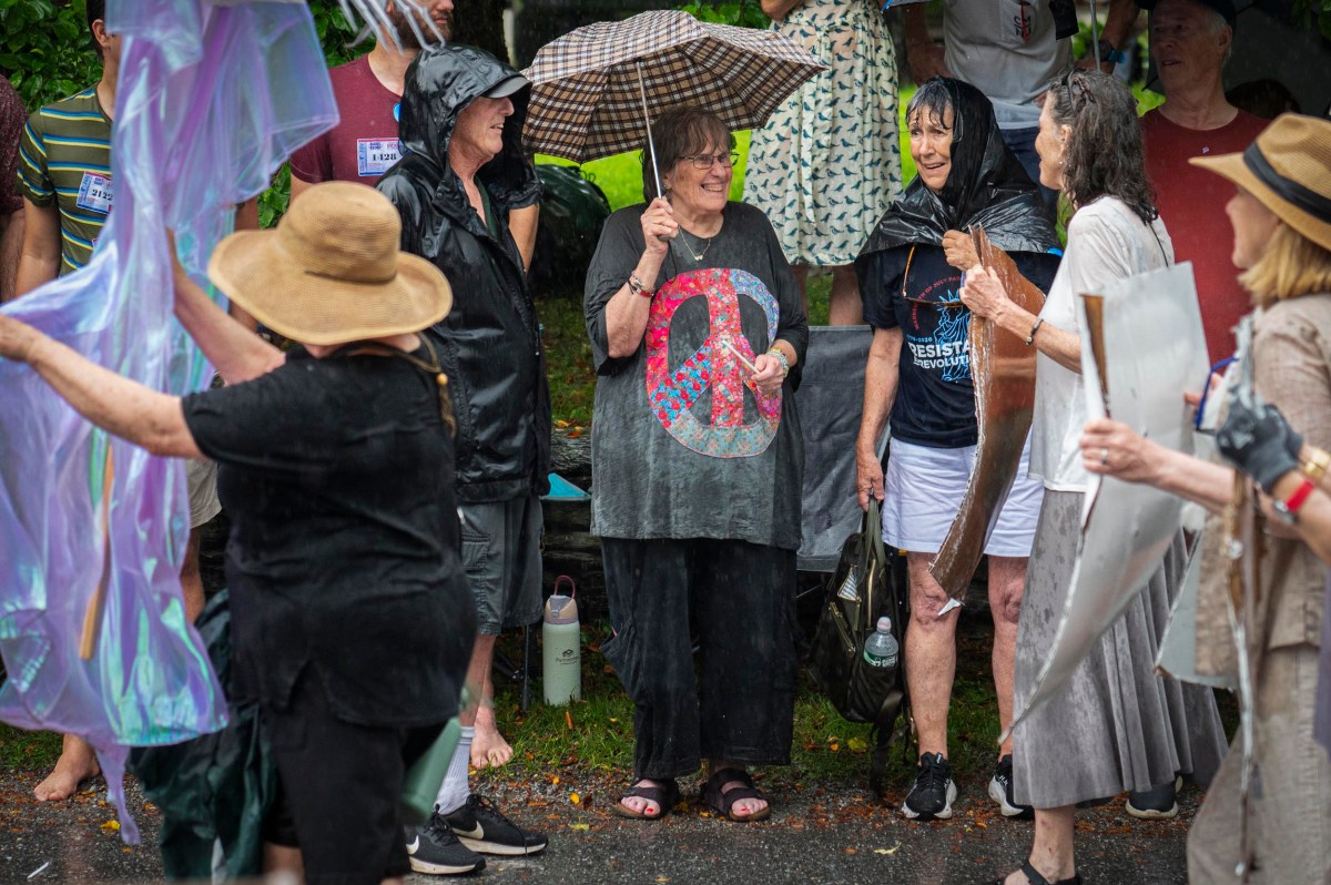 A group of people stand together outside in the rain, some holding umbrellas and wearing rain gear, while others interact and smile.