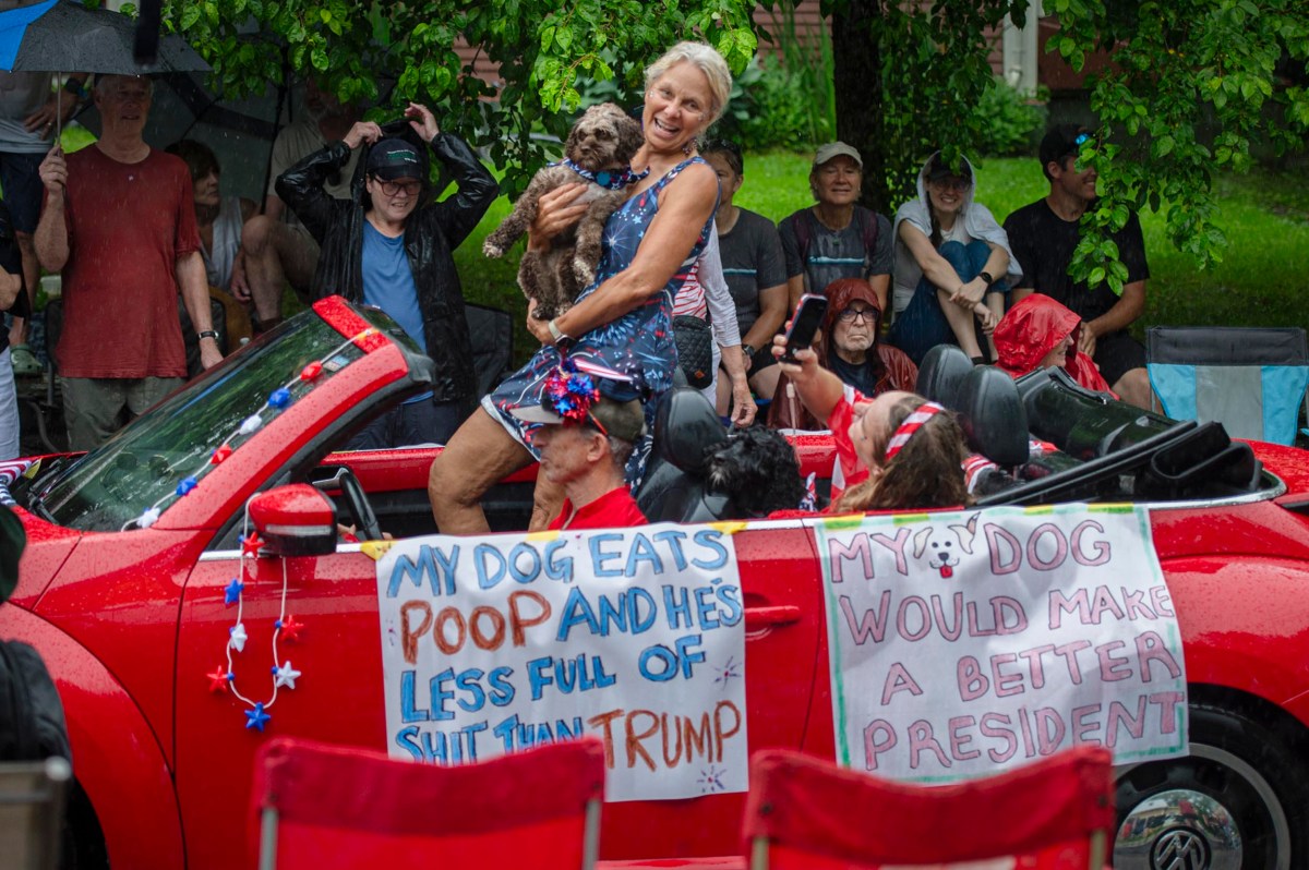 A woman holding a dog sits on the back of a red convertible in a parade; signs on the car make humorous political and dog-related statements. Spectators watch, some with umbrellas.