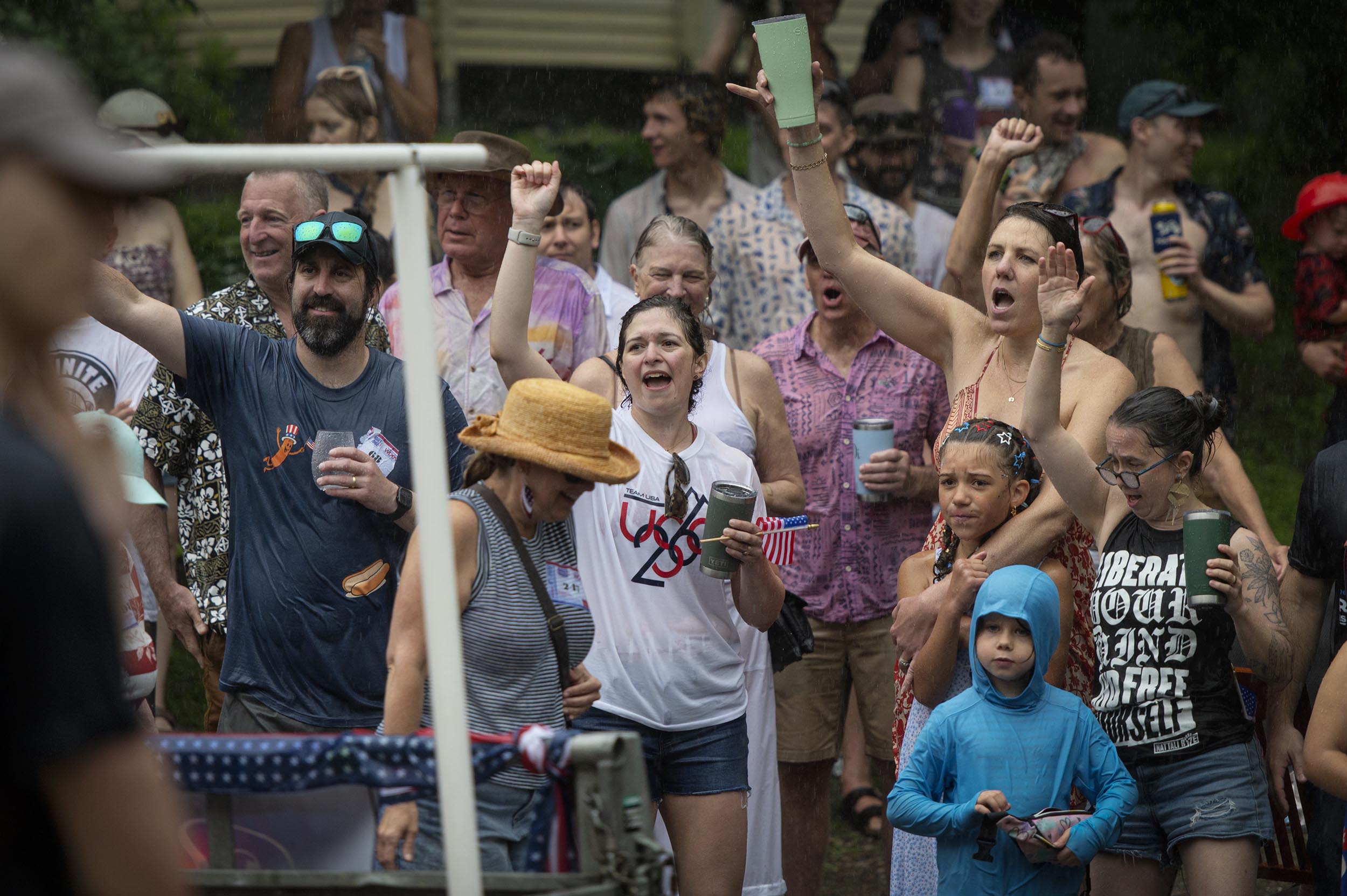 A group of people, some holding drinks and wearing casual clothes, cheer and raise their arms at an outdoor event in the rain.