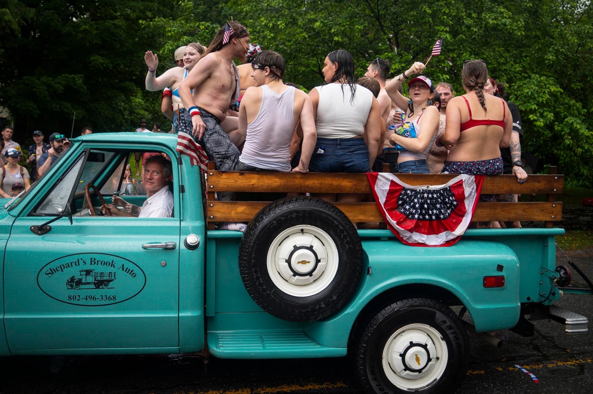 A group of people in swimsuits stand and sit in the bed of a turquoise pickup truck decorated with patriotic bunting during a parade on a rainy day.