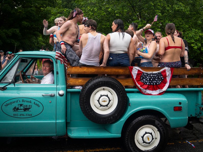 A group of people in swimsuits stand and sit in the bed of a turquoise pickup truck decorated with patriotic bunting during a parade on a rainy day.