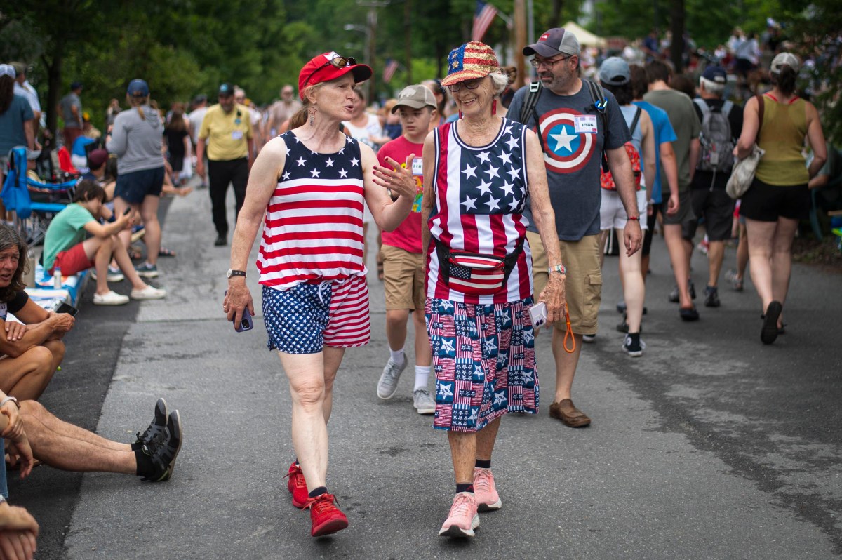 Two women dressed in American flag-themed outfits walk together at an outdoor event, surrounded by other people, some also wearing patriotic clothing.