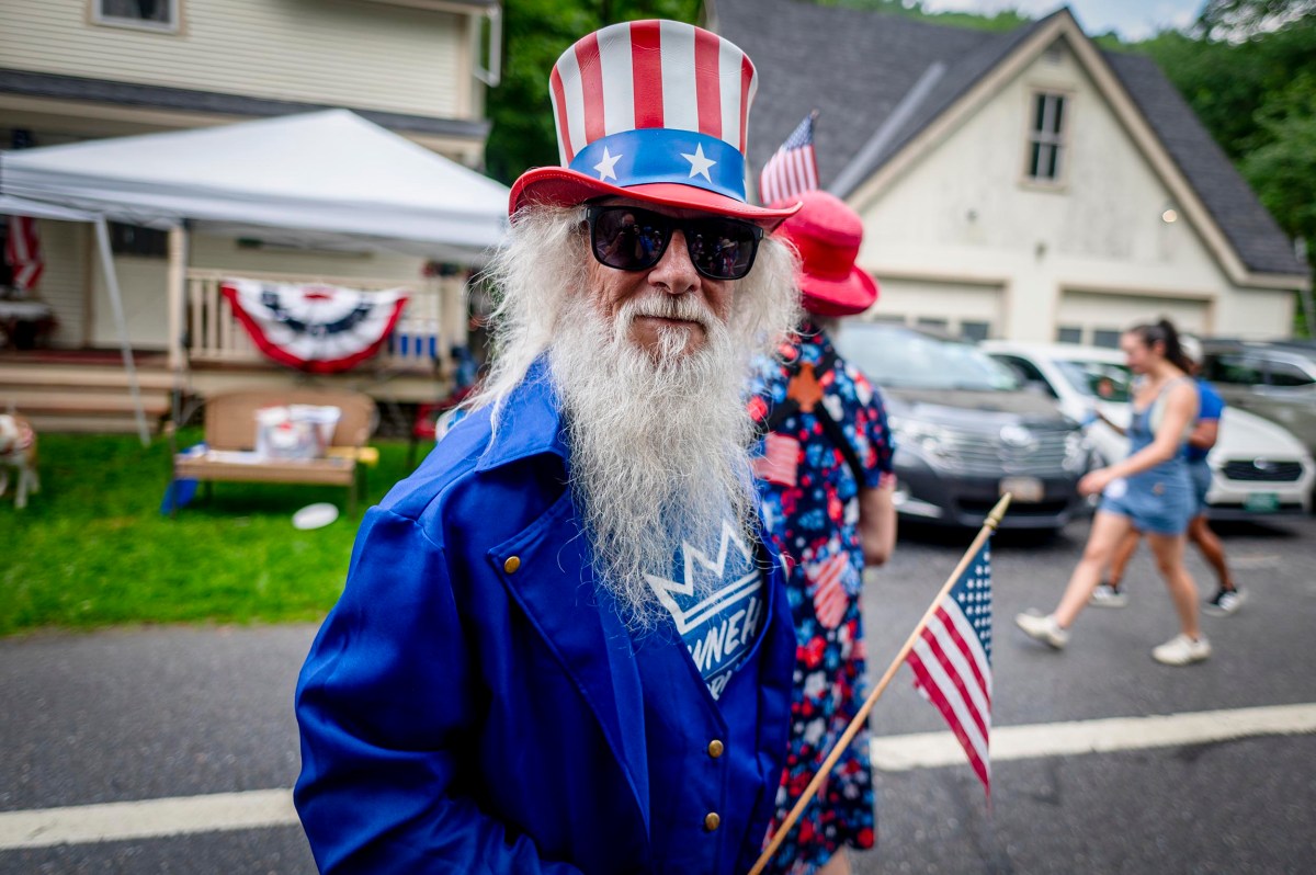 An older man with a long white beard wears a red, white, and blue Uncle Sam hat and jacket, holding an American flag at an outdoor event with people and houses in the background.
