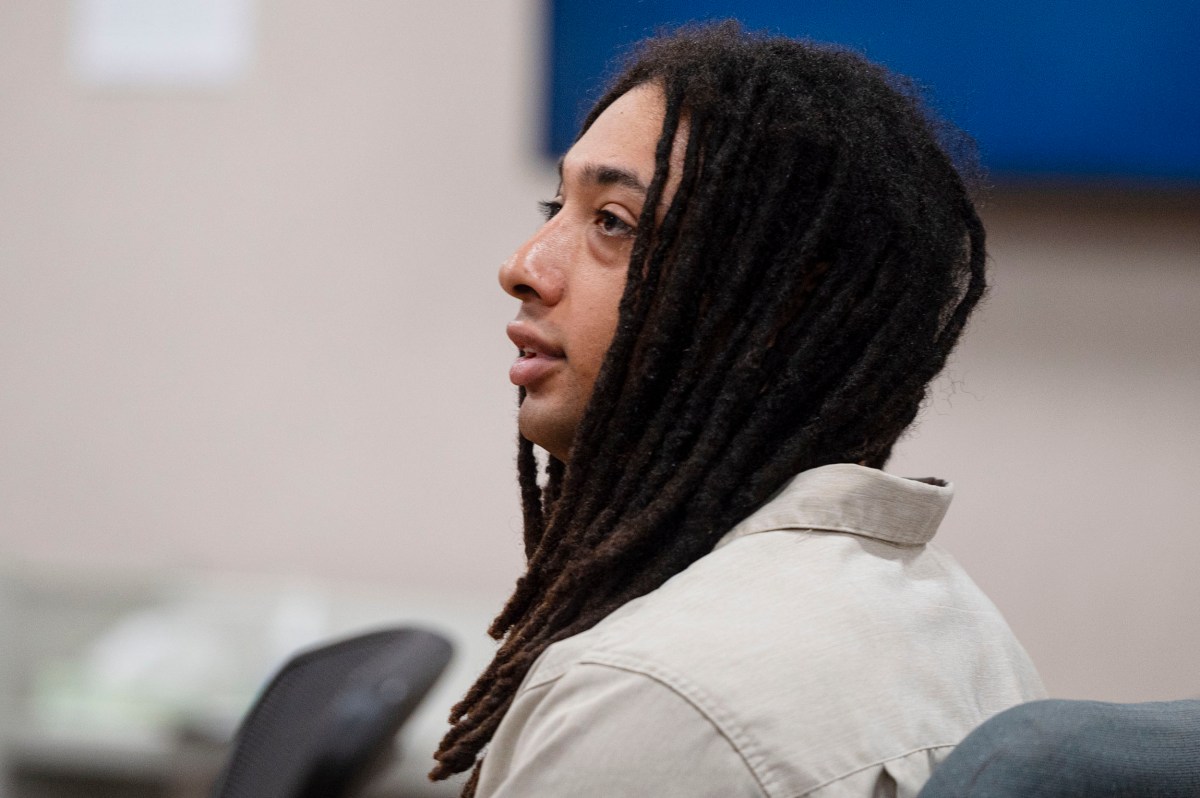 A person with long dreadlocks sits in a room, facing to the left. They wear a light-colored shirt and appear to be listening intently.