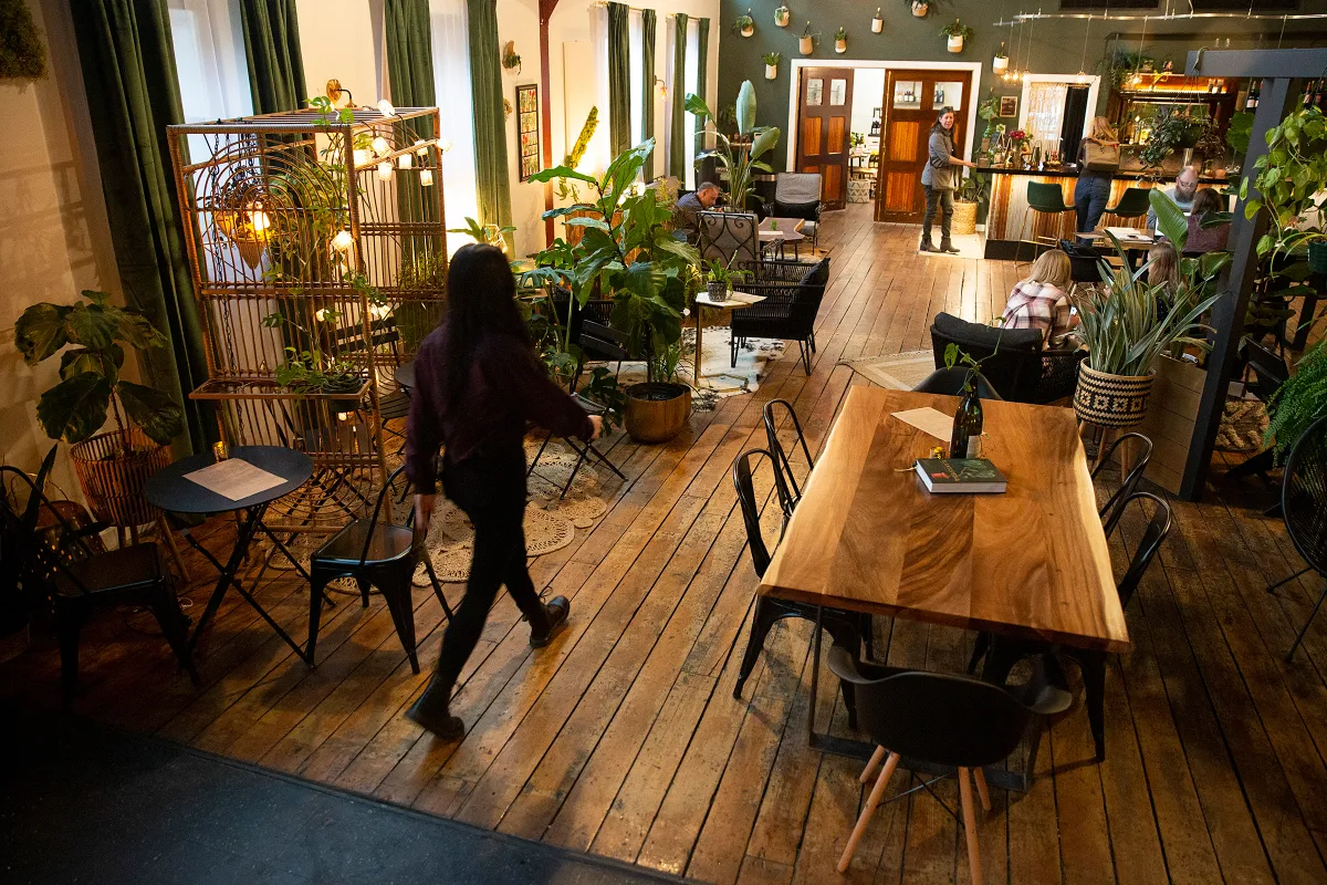 Spacious café with wooden floors, large plants, various tables and chairs, and several people sitting and walking inside a cozy, well-lit room.