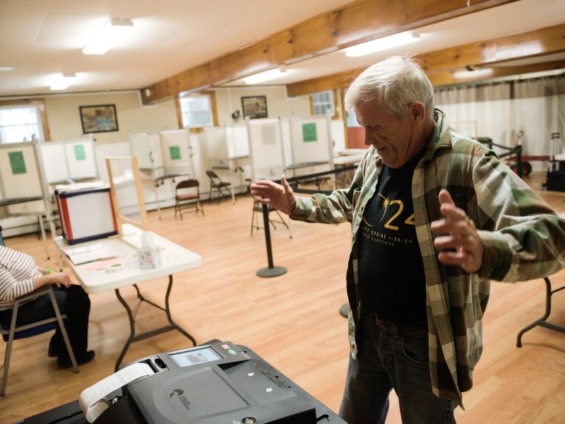 An older man stands with arms raised near a ballot machine in a polling station, while a woman sits at a table in the background.