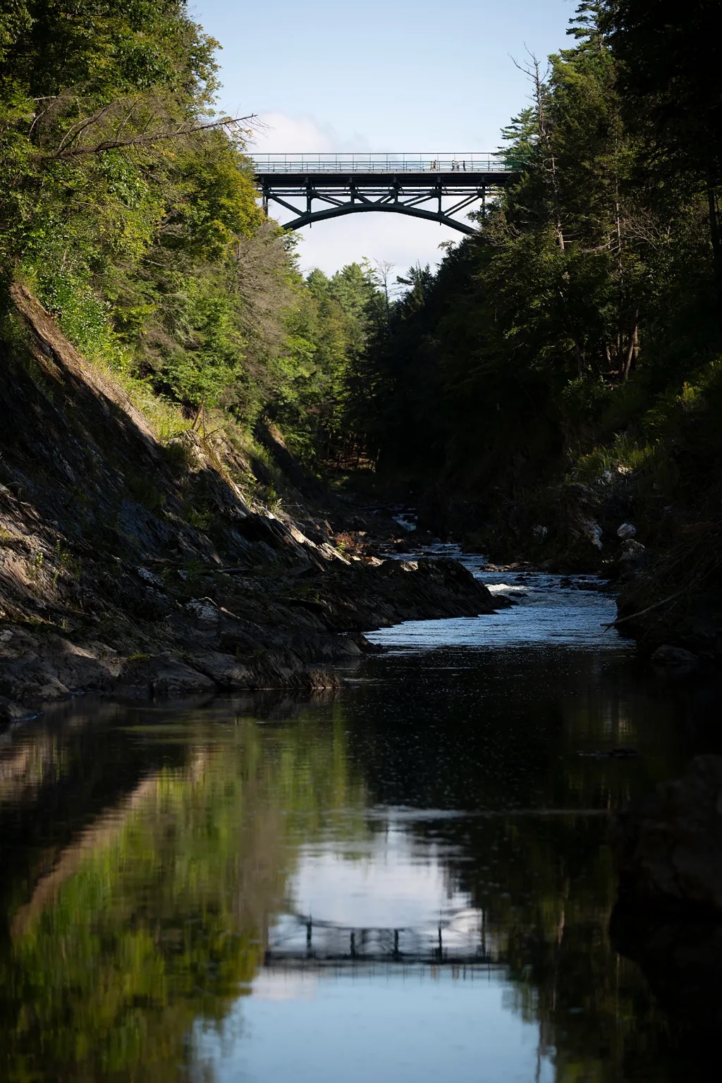 A metal bridge spans over a rocky creek surrounded by trees, with reflections of the bridge visible in the water below.