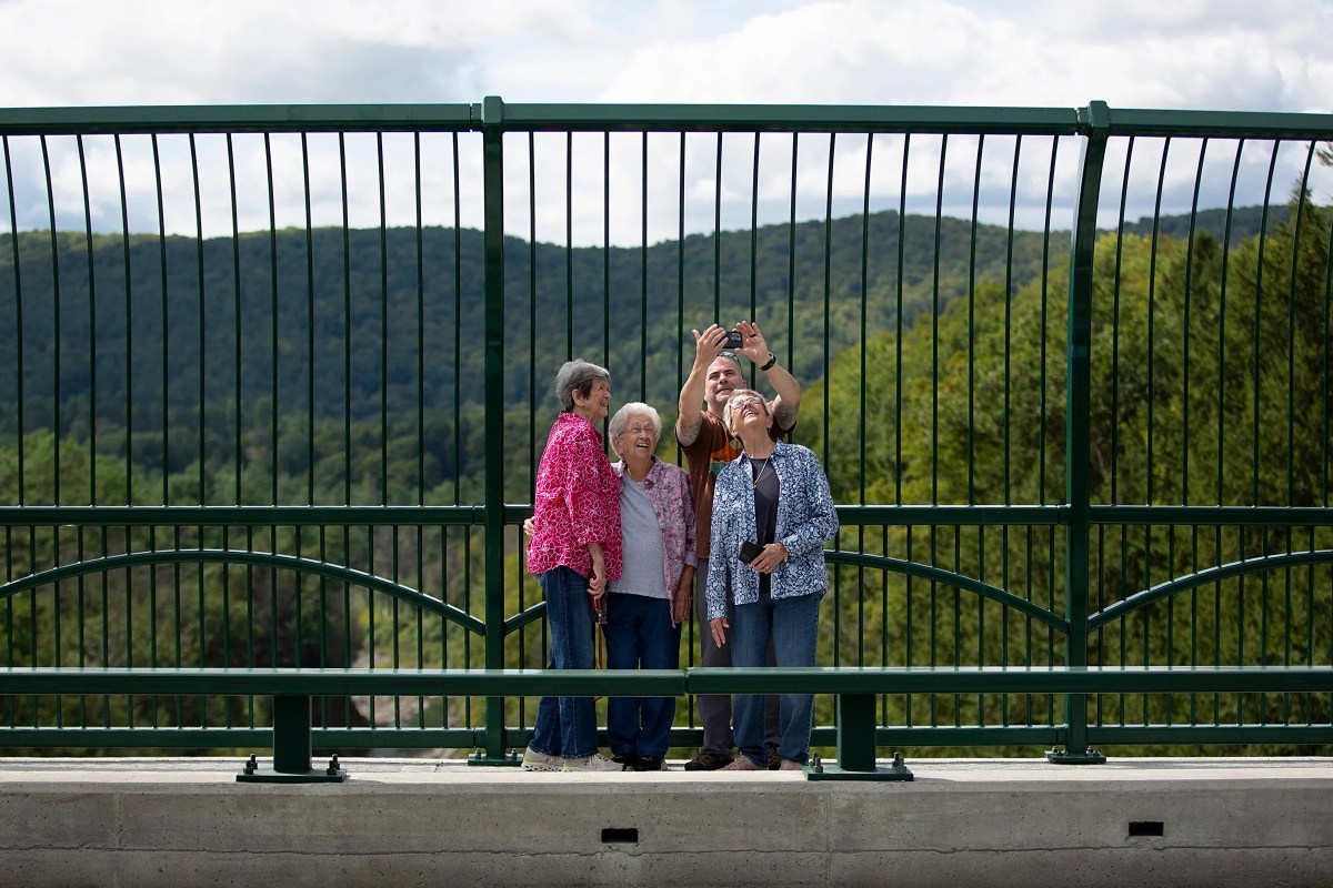 Four older adults stand together in front of a green fence on a bridge, posing for a selfie with trees and mountains in the background.