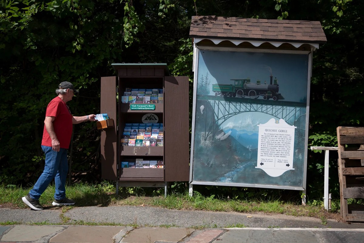A man in a red shirt and blue jeans takes a brochure from an outdoor display next to a large train-themed informational sign.
