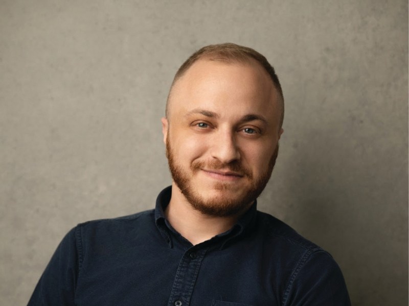 A person with short hair and a trimmed beard wearing a dark shirt is smiling slightly, seated in front of a plain, light gray background.