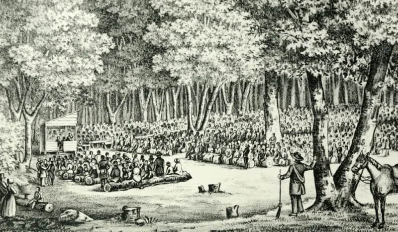 A large group of people gathers in a forest clearing, listening to a speaker on an outdoor platform, with trees and more onlookers in the background.