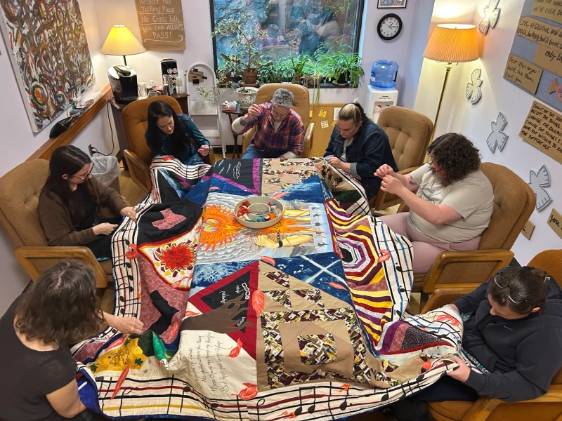Eight people sit in a circle, working together on a large, colorful quilt in a cozy, well-lit room decorated with paintings, posters, and comfortable furniture.