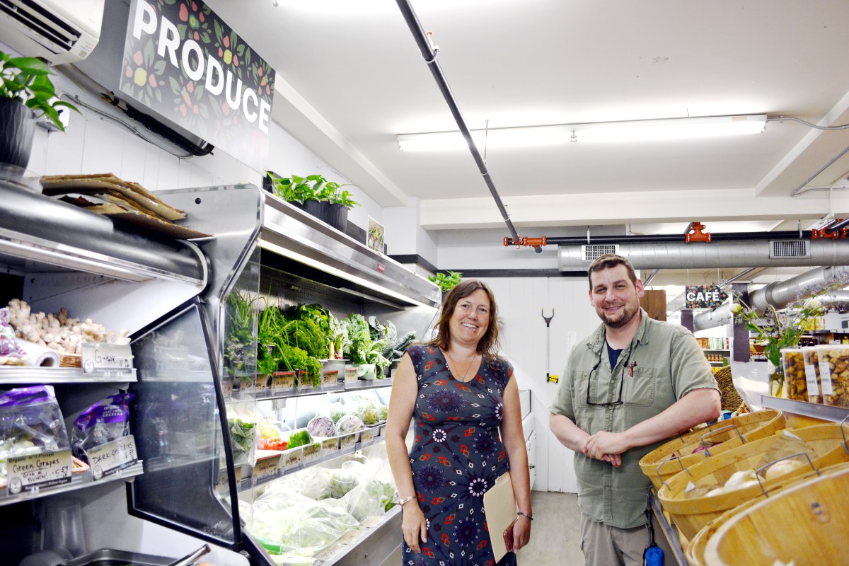 Two people stand and smile near shelves of fresh produce and bread in a grocery store with a “Produce” sign overhead.