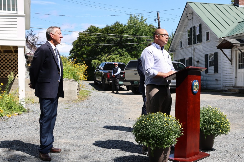 A man stands at a podium speaking outdoors while another man stands nearby; two people and parked vehicles are visible in the background on a sunny day.