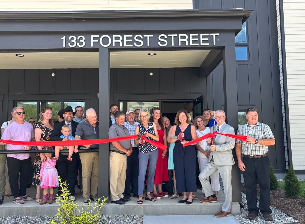 A group of people stand in front of a building labeled “133 Forest Street” holding a large red ribbon for a ribbon-cutting ceremony.