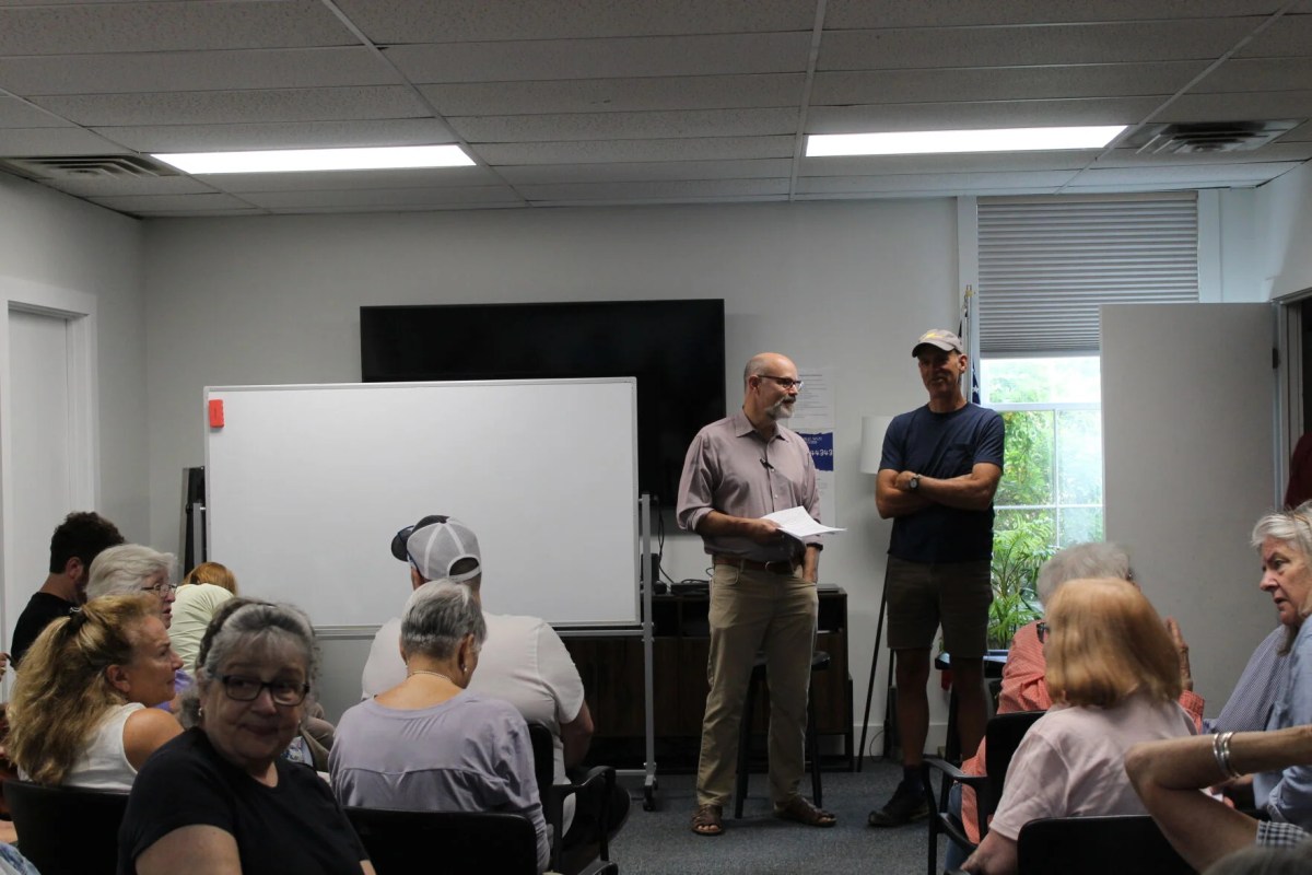 A group of people sit facing two men standing in front of a whiteboard in a room with fluorescent lighting and a television mounted on the wall.