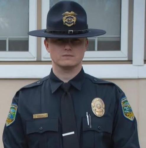 Police officer in a dark uniform and hat stands in front of a building, facing forward with a neutral expression.