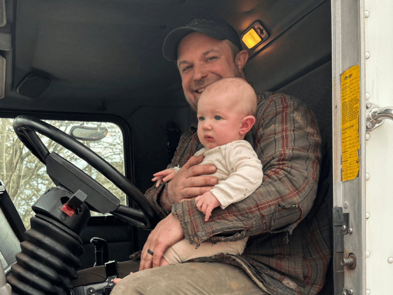 A man in a plaid shirt holds a baby while sitting in the driver’s seat of a truck. The man is smiling, and the baby looks forward with a curious expression.