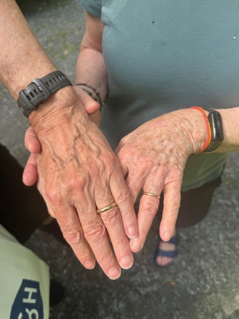 Two people stand next to each other, showing their hands with wedding rings and wristwatches. They are outdoors on a gravel surface.